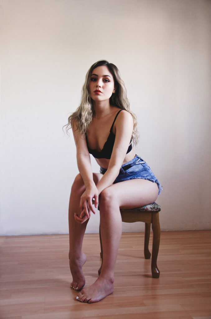 Darian Rojas poses elegantly on a wooden stool against a plain wall wearing a black bralette and denim shorts with her long blonde hair cascading down her shoulders.